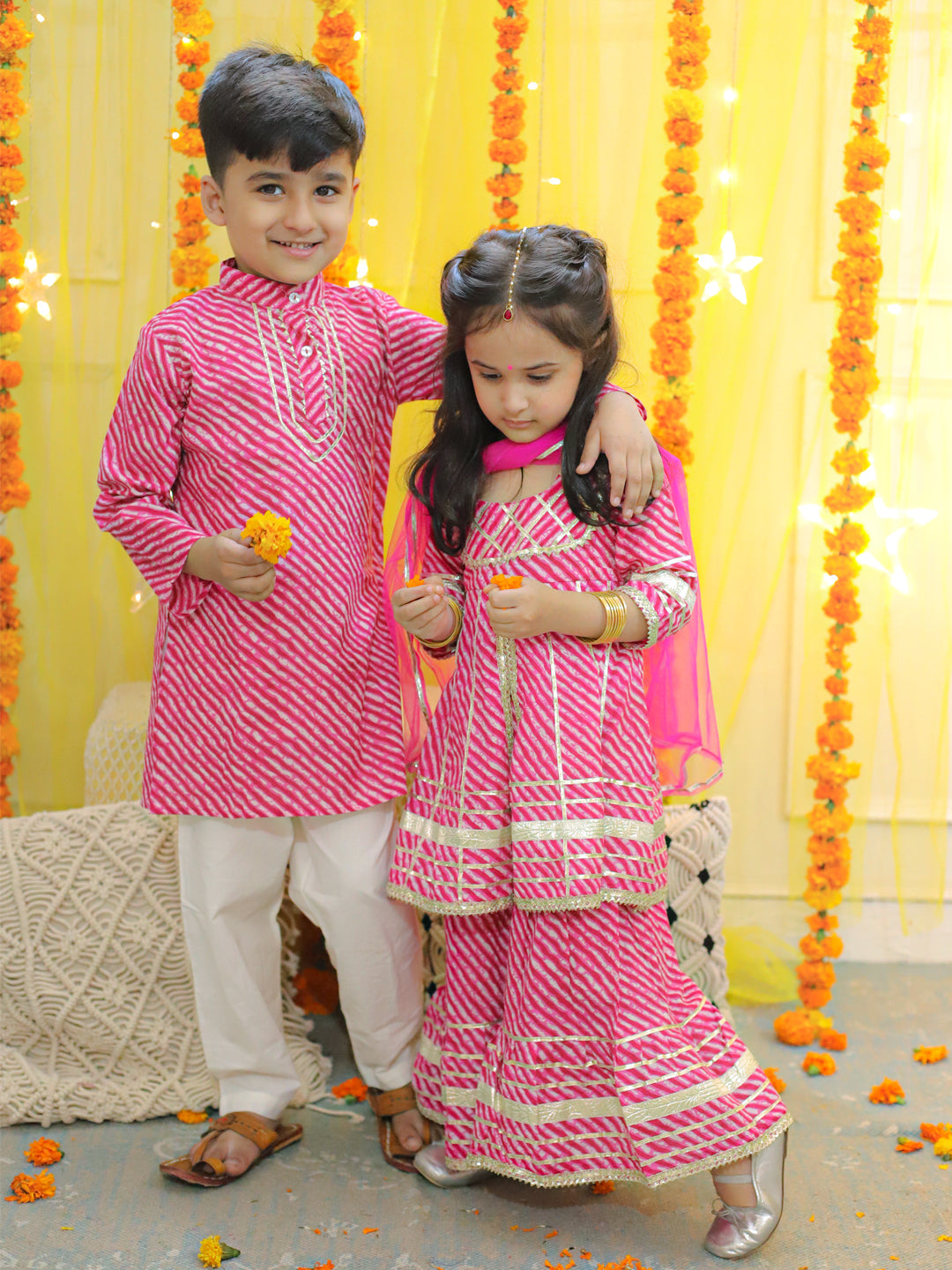 Two children, a boy in a pink shirt with a pattern and a girl in a pink dress with a dupatta, standing next to each other with a background of yellow decorations and flowers.