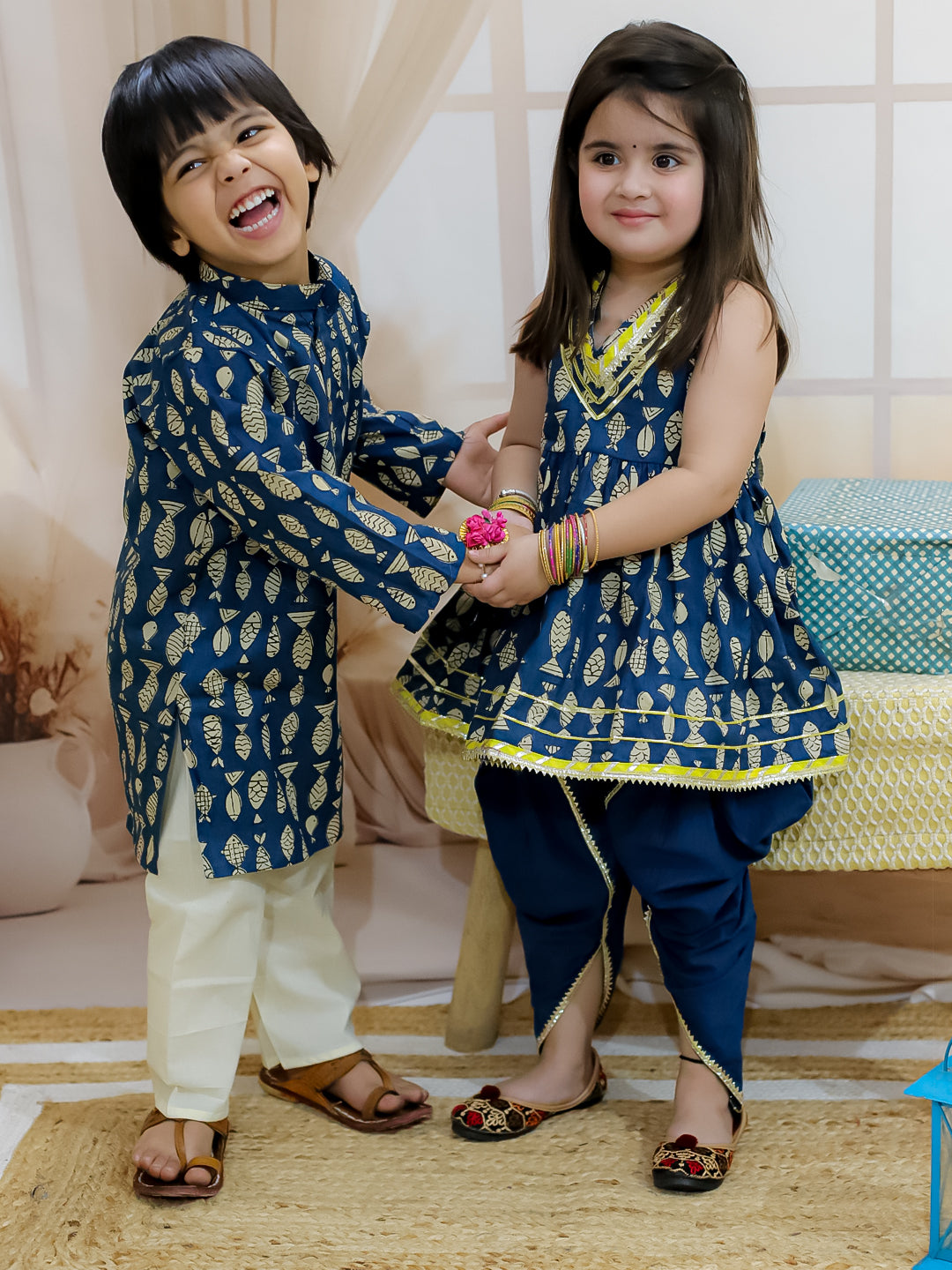 A child in a blue kurta pajama set with a fish print interacting with another child who is wearing a matching blue dress with a halter neck top. Both are smiling and posing for the camera.