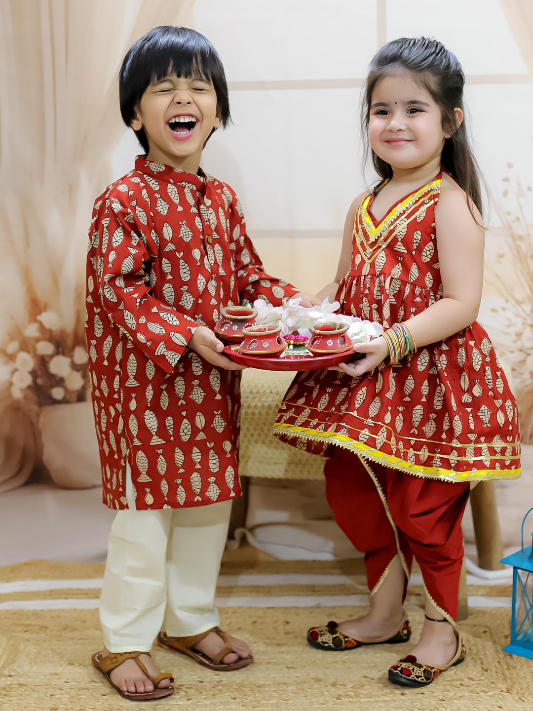 Two children, a boy and a girl, wearing matching maroon kurta pajama sets with fish prints and halter neck tops, holding hands and smiling.