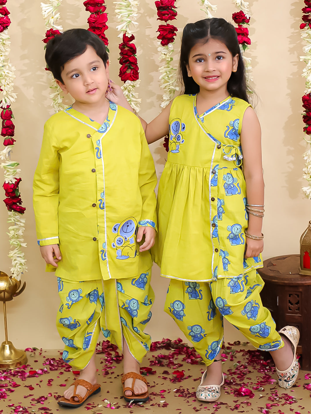 Two children in matching yellow traditional outfits with blue patterns, standing against a decorated background.